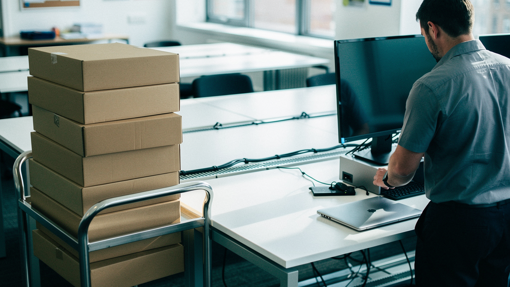 A bright UK open-plan office on a weekday morning, a small trolley stacked with unopened laptop boxes beside a row of desks, an IT technician unpacking one laptop and placing it next to a docking stat