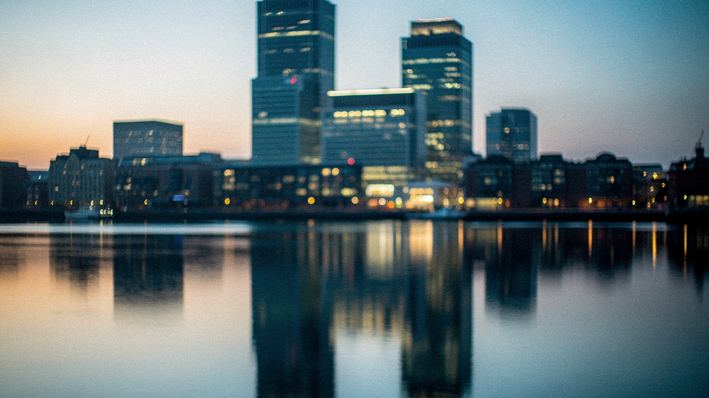 A row of glass office towers in the City of London at dusk, upper floors lit from within, an empty wet plaza in the foreground reflecting the lights, two distant pedestrian silhouettes crossing far be
