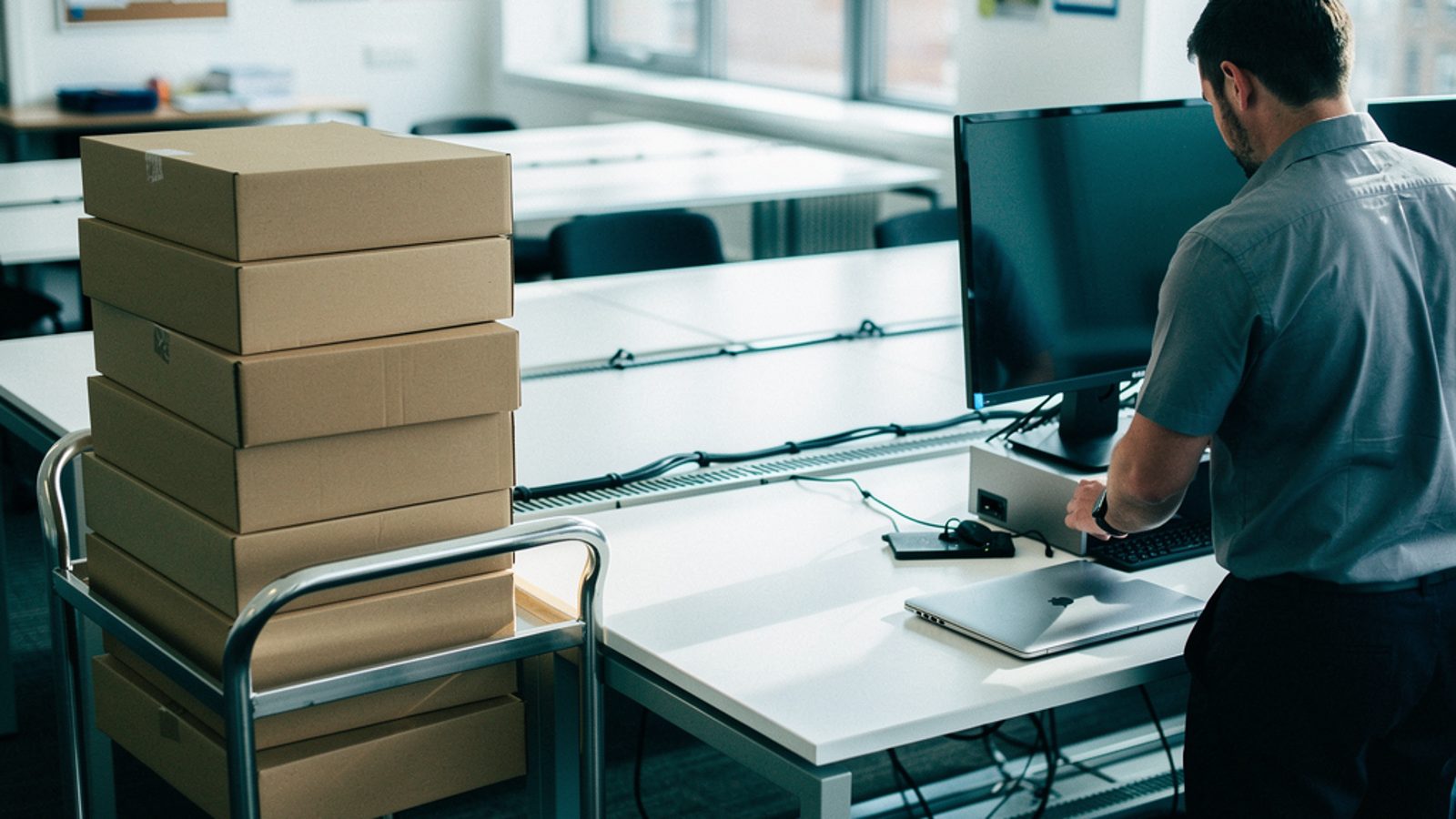 A bright UK open-plan office on a weekday morning, a small trolley stacked with unopened laptop boxes beside a row of desks, an IT technician unpacking one laptop and placing it next to a docking stat