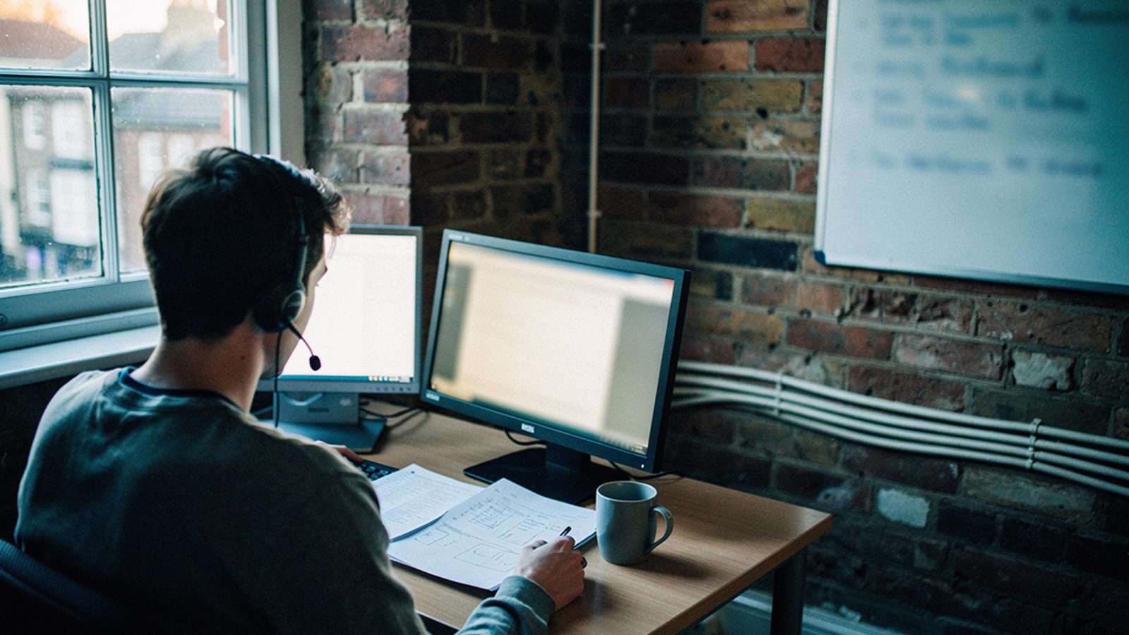 A small IT support office in a converted terrace building in a UK town, early morning light through a sash window. A technician is seen from behind at a desk, headset on, papers and a coffee mug nearb