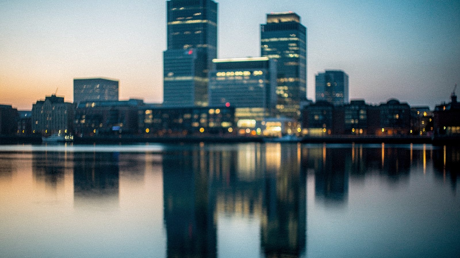 A row of glass office towers in the City of London at dusk, upper floors lit from within, an empty wet plaza in the foreground reflecting the lights, two distant pedestrian silhouettes crossing far be