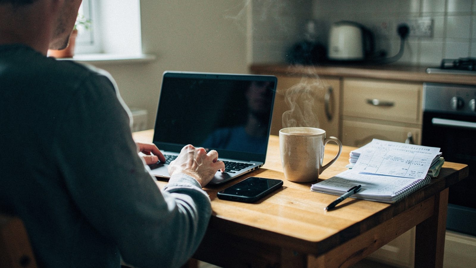 A UK sole trader working at a small kitchen table in a terraced house, laptop open beside a phone and a mug of tea, morning light through a nearby window, a stack of invoices and a notebook nearby, no