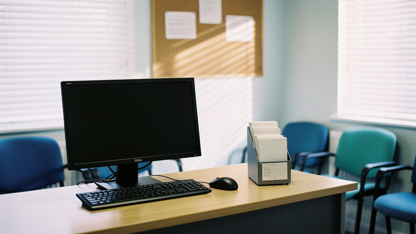 The quiet reception area of a small UK GP surgery in early morning light, a switched-off desktop monitor on the front desk, a patient appointment card rack and a wall noticeboard with pinned notices n