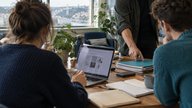 A small team in a bright meeting room at a Bristol co-working space, comparing Google Workspace plans on an open laptop while a colleague points to a stack of project folders and an external drive. Fr