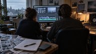 Inside a small British film-editing studio on an overcast afternoon, a producer and sound editor seen from behind review a video timeline beside a mixing console. Printed photographic contact sheets,
