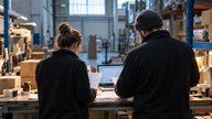A small British wholesale warehouse during an early morning operations review, photographed from behind two staff members beside a packing bench. They examine anonymised order printouts and a laptop s