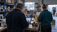 A small British wholesale warehouse in late afternoon, photographed over the shoulder of a sales supervisor reviewing a proposed customer quotation on a tablet beside a packing bench. A colleague wait