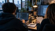 A small British software studio during a late-afternoon code review, photographed over the shoulders of two developers with faces outside the frame. One laptop shows an indistinct code editor while a
