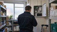 A maintenance engineer seen from behind inspecting an open security alarm cabinet in the stockroom of a small British high-street shop. A compact cellular communicator, antenna cable and neatly labell
