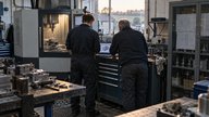A realistic editorial photograph inside a small British precision engineering workshop in late afternoon. Two staff members, seen from behind with no identifiable faces, review a laptop beside a conne