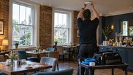 A realistic editorial photograph inside a small British hotel breakfast room during early morning setup. A technician seen from behind checks a ceiling-mounted wireless access point while an open equi