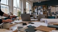 A small British architecture studio during a morning equipment pilot, with two open laptops and three different smartphones arranged on a shared project table beside rolled building plans. An IT techn