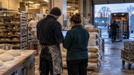 A realistic editorial photograph inside a British independent wholesale bakery at dawn, showing a delivery supervisor and bookkeeper from behind checking a tablet beside stacked flour sacks and stainl
