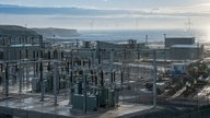 A realistic editorial photograph of an industrial site on the Northumberland coast, with a high-voltage substation in the foreground, low industrial buildings beyond and offshore wind turbines visible