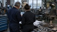 Inside a small British metalworking factory on an overcast autumn morning, an owner and technician seen from behind compare a tablet showing an unbranded production interface with paper job sheets bes