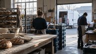 An early-morning scene inside a small British wholesale bakery, where an owner seen from behind checks a tablet beside stacked delivery crates while a colleague sorts dispatch slips at a loading hatch