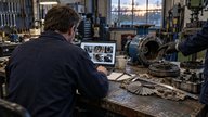 A surveyor seen from behind inside a small British engineering workshop, reviewing inspection photographs on a slim laptop beside a dismantled pump and neatly arranged measuring tools. A colleague's g