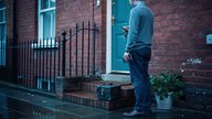 A UK landlord standing at the front door of a let terraced house in early evening light, holding a bunch of house keys and a phone, a small toolbox and a potted plant at their feet, brick steps and a