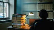 A small UK solicitors' office in the late afternoon, a practice manager sitting at a desk with a closed laptop and a stack of paper folders, a frosted glass partition and rows of box files visible beh