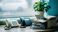 A small UK office reception desk at morning light, an old cream analogue telephone receiver resting beside a new black VoIP desk handset and a half-unpacked cardboard box with coiled cables, a wall ca