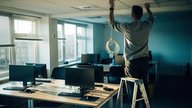 A small UK open-plan office in late-afternoon light, an IT technician viewed from behind on a step ladder fitting a compact white ceiling-mounted wireless access point, a coil of thin ethernet cable i
