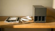 A tidy home-office shelf at eye level holding a small dark grey desktop NAS enclosure with its front drive bays open, one bare 3.5 inch hard drive resting on a soft cloth beside it, a coiled short eth