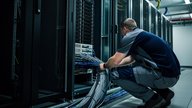 A UK data centre server room technician in dark workwear, seen from behind, standing at an open server rack inspecting cabling by torchlight. Rows of unlit rack cabinets recede into shadow. Cool blue-