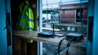 A portable cellular router with a stub antenna sitting on a plywood shelf inside a UK construction site cabin, cables running to a small switch, high-vis jacket hanging on a hook nearby, seen through
