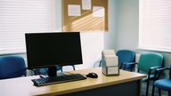 The quiet reception area of a small UK GP surgery in early morning light, a switched-off desktop monitor on the front desk, a patient appointment card rack and a wall noticeboard with pinned notices n
