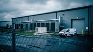 A colocation data centre building exterior on a business park in England at early morning, chain-link perimeter fencing, external cooling units and a standby generator along the building's side wall,