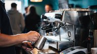 A small independent UK coffee shop counter at mid-morning, barista's hands steaming milk, a compact card payment terminal sitting on the counter beside a till, warm pendant lighting, condensation on t