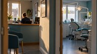 A realistic editorial photograph of a small UK dental practice reception and single treatment room seen from the corridor, warm morning light through a front window, a practice manager reviewing a clo