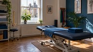 A small private physiotherapy clinic treatment room in a UK high street building, mid-morning light through a sash window, a height-adjustable treatment couch with folded blue towels, a set of resista
