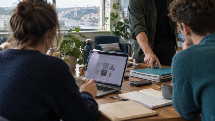 A small team in a bright meeting room at a Bristol co-working space, comparing Google Workspace plans on an open laptop while a colleague points to a stack of project folders and an external drive. Fr