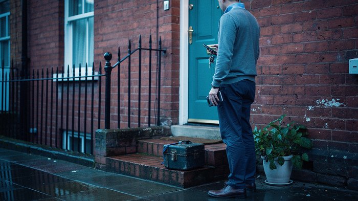 A UK landlord standing at the front door of a let terraced house in early evening light, holding a bunch of house keys and a phone, a small toolbox and a potted plant at their feet, brick steps and a
