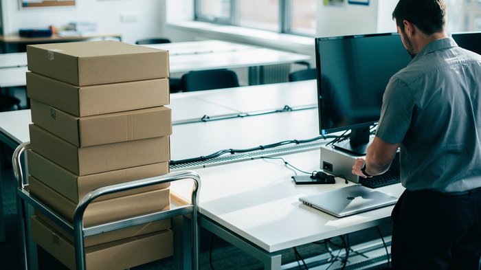 A bright UK open-plan office on a weekday morning, a small trolley stacked with unopened laptop boxes beside a row of desks, an IT technician unpacking one laptop and placing it next to a docking stat