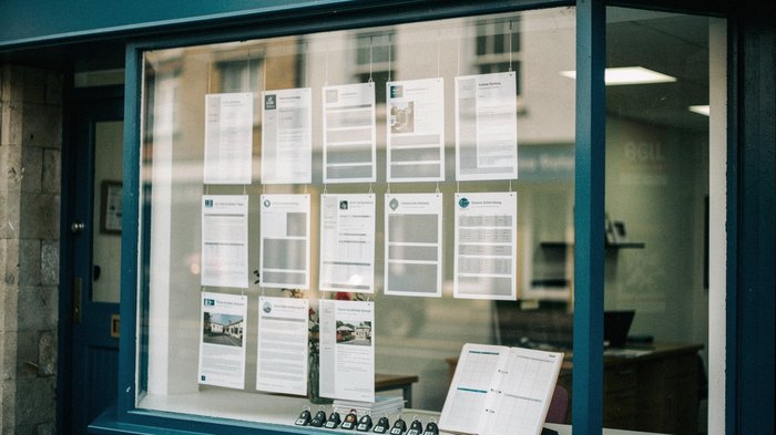 A small independent UK estate agency's high street shopfront in an English market town at opening time, printed property particulars displayed behind the window glass, a set of labelled house keys and