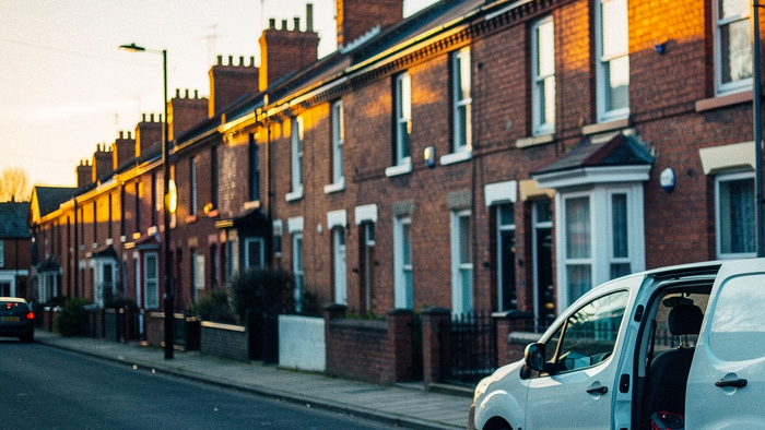 A red-brick Victorian terraced street in a UK town at golden hour. A small tradesperson's van is parked outside one house with its side door open, a folder of paperwork resting on the seat. A leaning