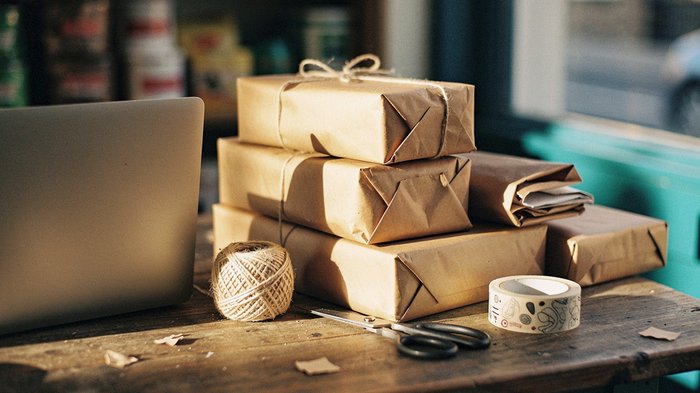 A small UK independent shop's packing table mid-afternoon, brown-paper parcels stacked and half-wrapped, a laptop closed to one side, string and a roll of branded tape, warm low autumn light through a
