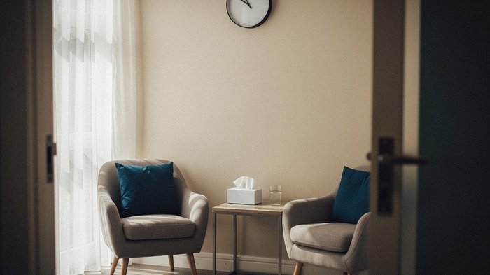 A quiet, softly lit UK counselling room seen from the doorway: two armchairs angled toward each other, a low side table with a box of tissues and a glass of water, a wall clock, soft daylight through