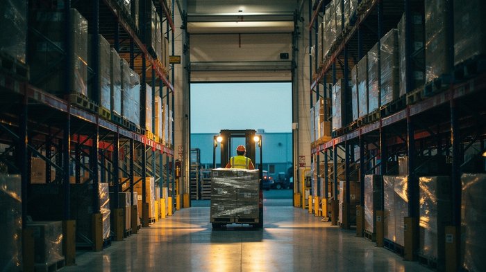 A UK third-party logistics warehouse at dusk, tall pallet racking stretching into the distance, a forklift carrying a wrapped pallet between two aisles, overhead sodium lighting warming the concrete f