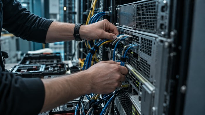 Inside a UK enterprise equipment room in the early evening, a technician's hands connect network cables to a single rack-mounted database server beside an open equipment case. Close framing shows cabl