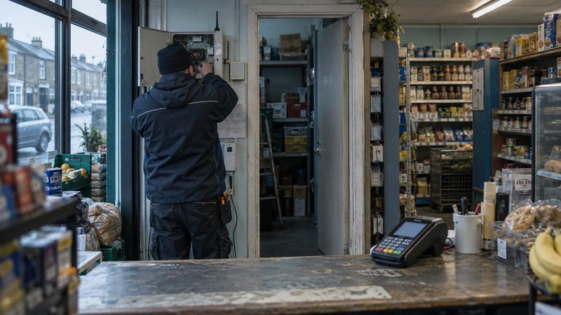 Inside a small British corner shop before opening on an overcast morning, an engineer seen from behind inspects an open alarm communications enclosure beside the stockroom doorway. A portable card ter